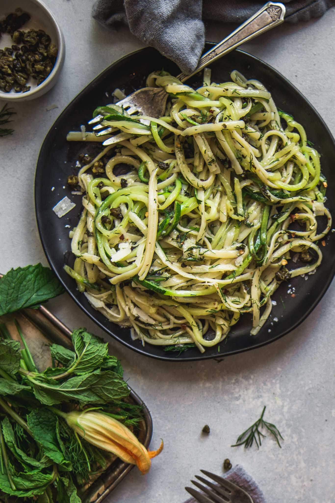 Overhead shot of dish of zucchini noodles with fresh herbs on a black plate.