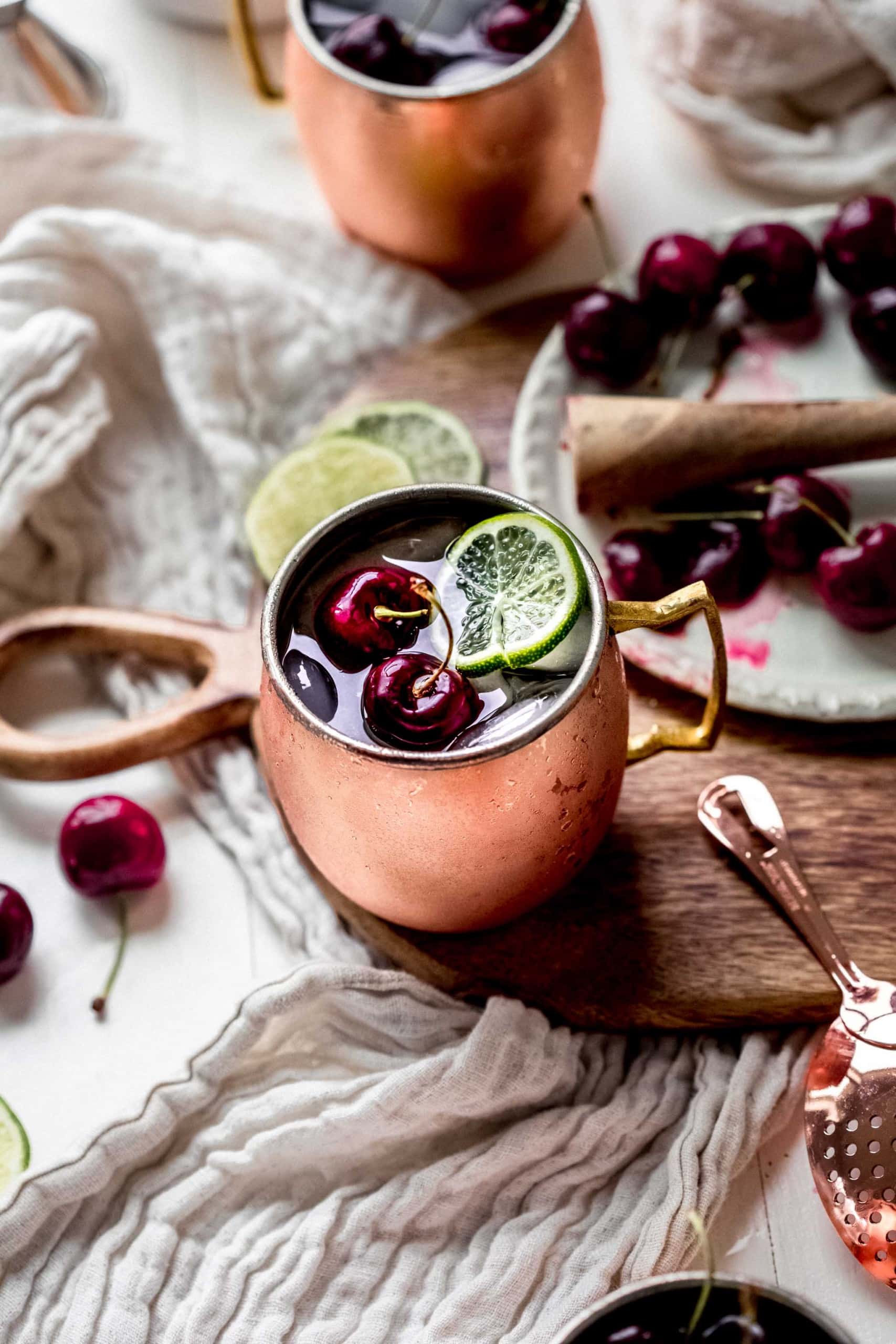 Two cherry moscow mule cocktails on counter next to pitted cherries and cocktail strainer.