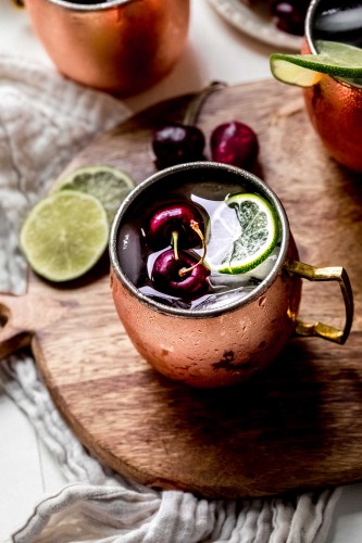 Two cherry moscow mule cocktails on counter next to pitted cherries and cocktail strainer.