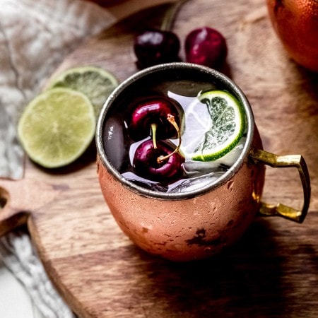 Two cherry moscow mule cocktails on counter next to pitted cherries and cocktail strainer.