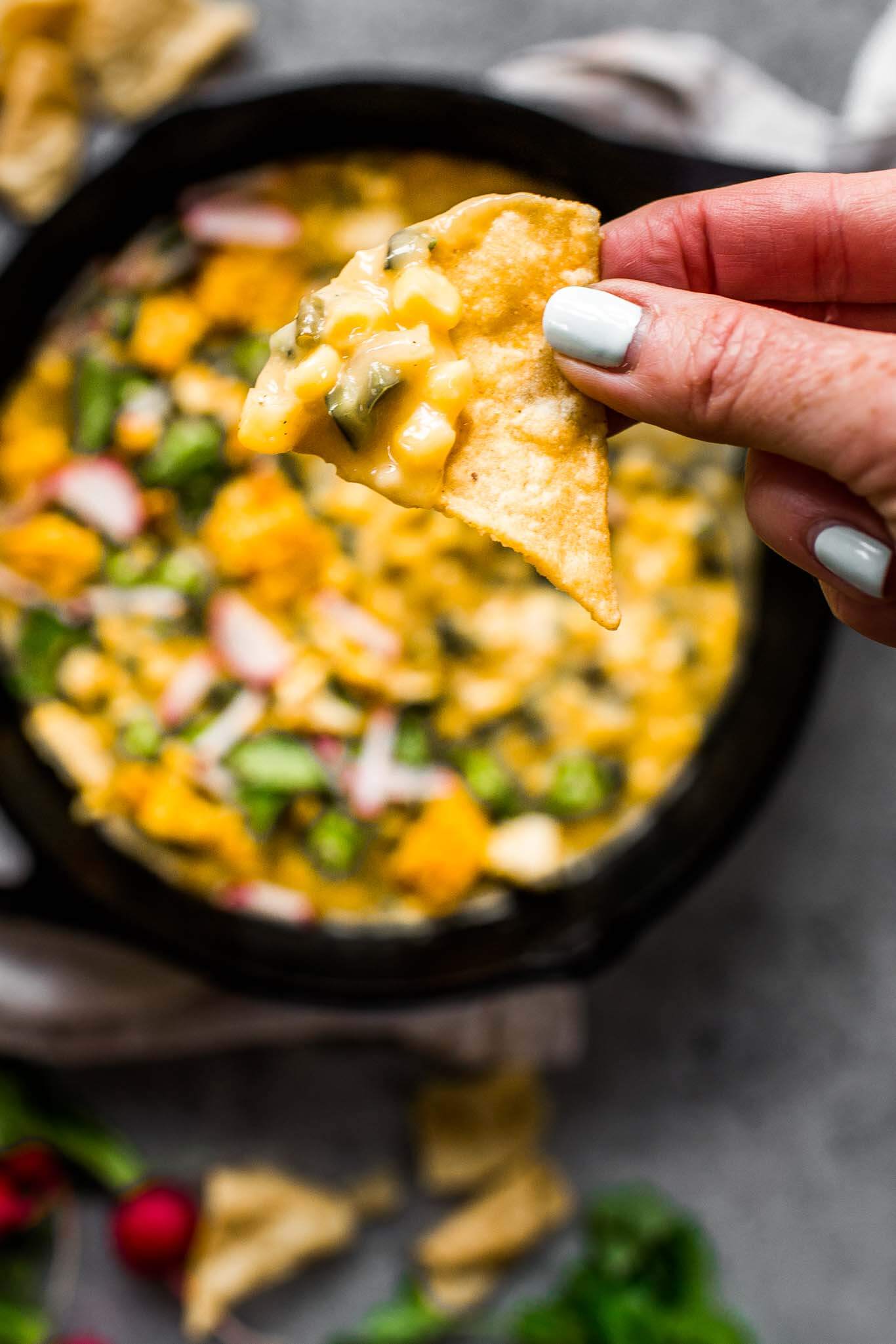 Hand holding tortilla chip dipped into corn queso dip