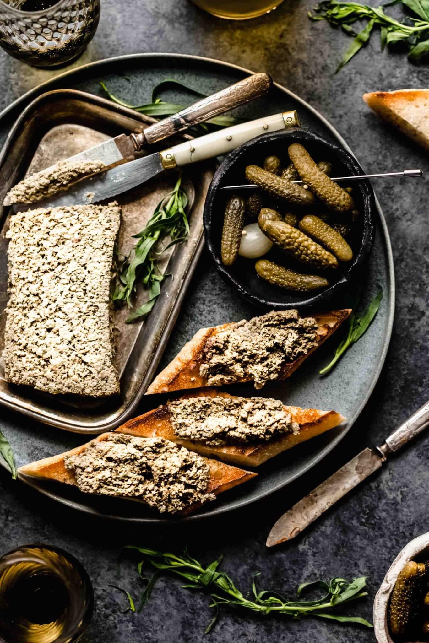 Mushroom pate on dark grey plate next to crostini and bowl of gherkins.