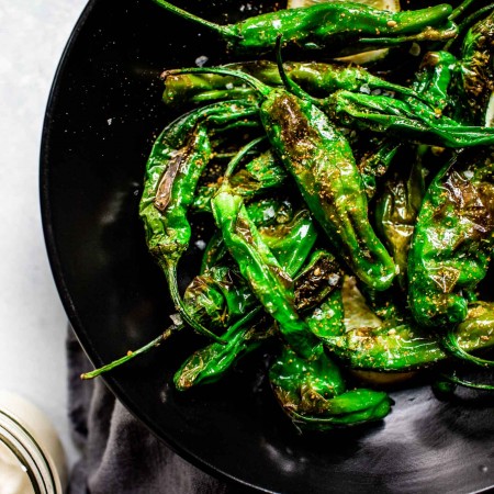 Blistered shishito peppers served on a black plate with lemon wedges.