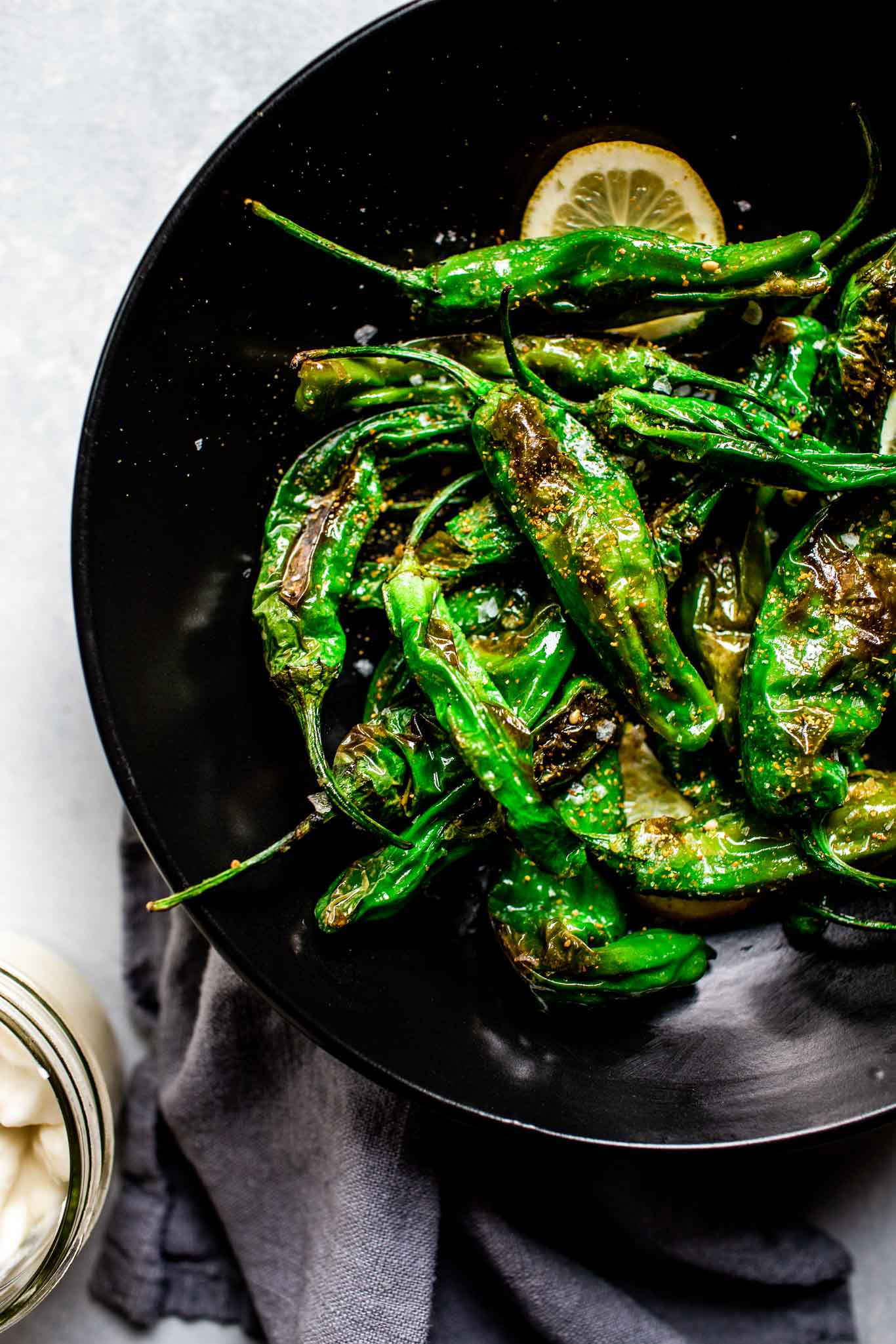 Blistered shishito peppers served on a black plate with lemon wedges.