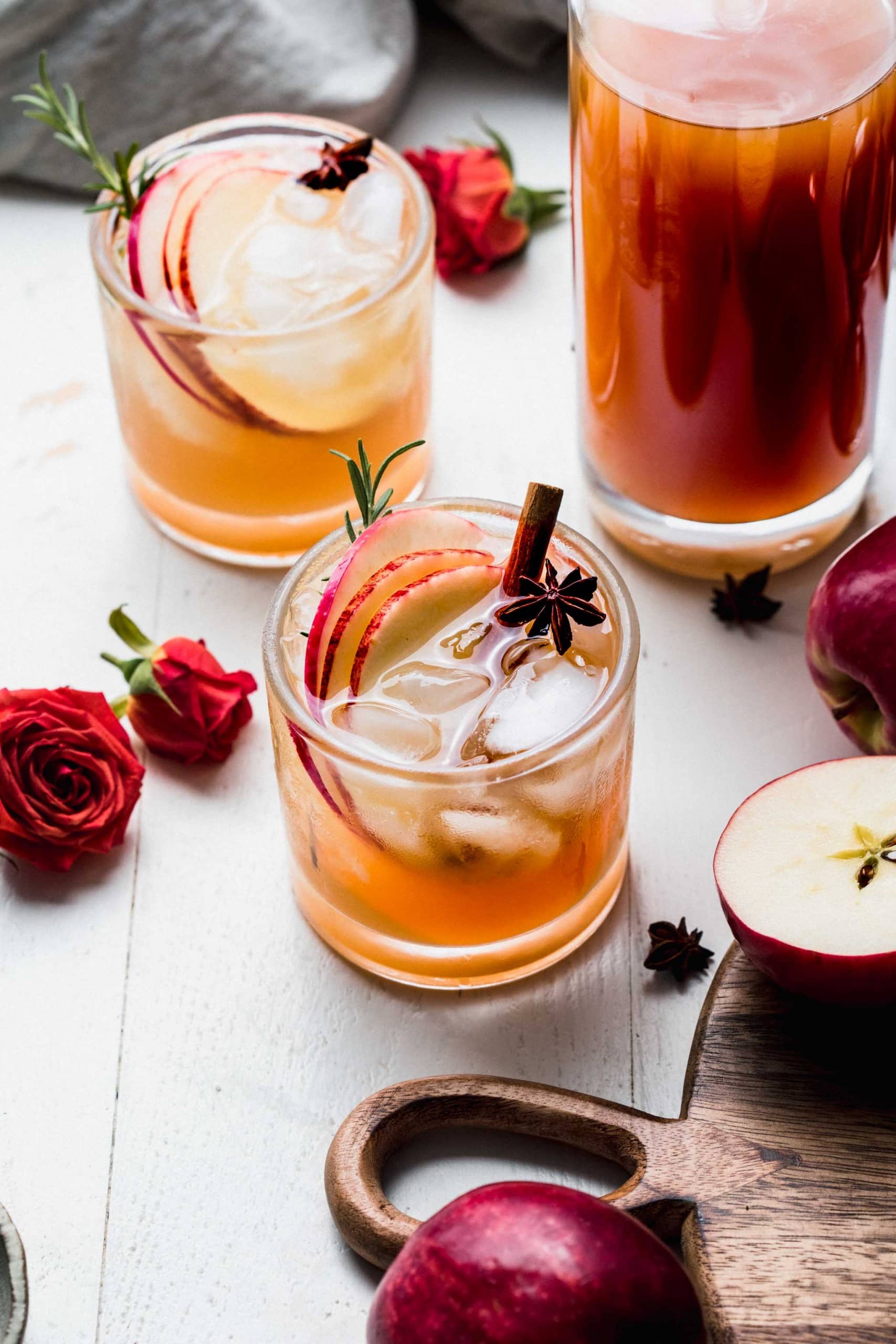 Side view of two apple cider cocktails next to jug of cider and red roses.