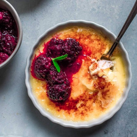 Overhead shot of creme brulee topped with roasted blackberries and mint sprig with spoon breaking into it.