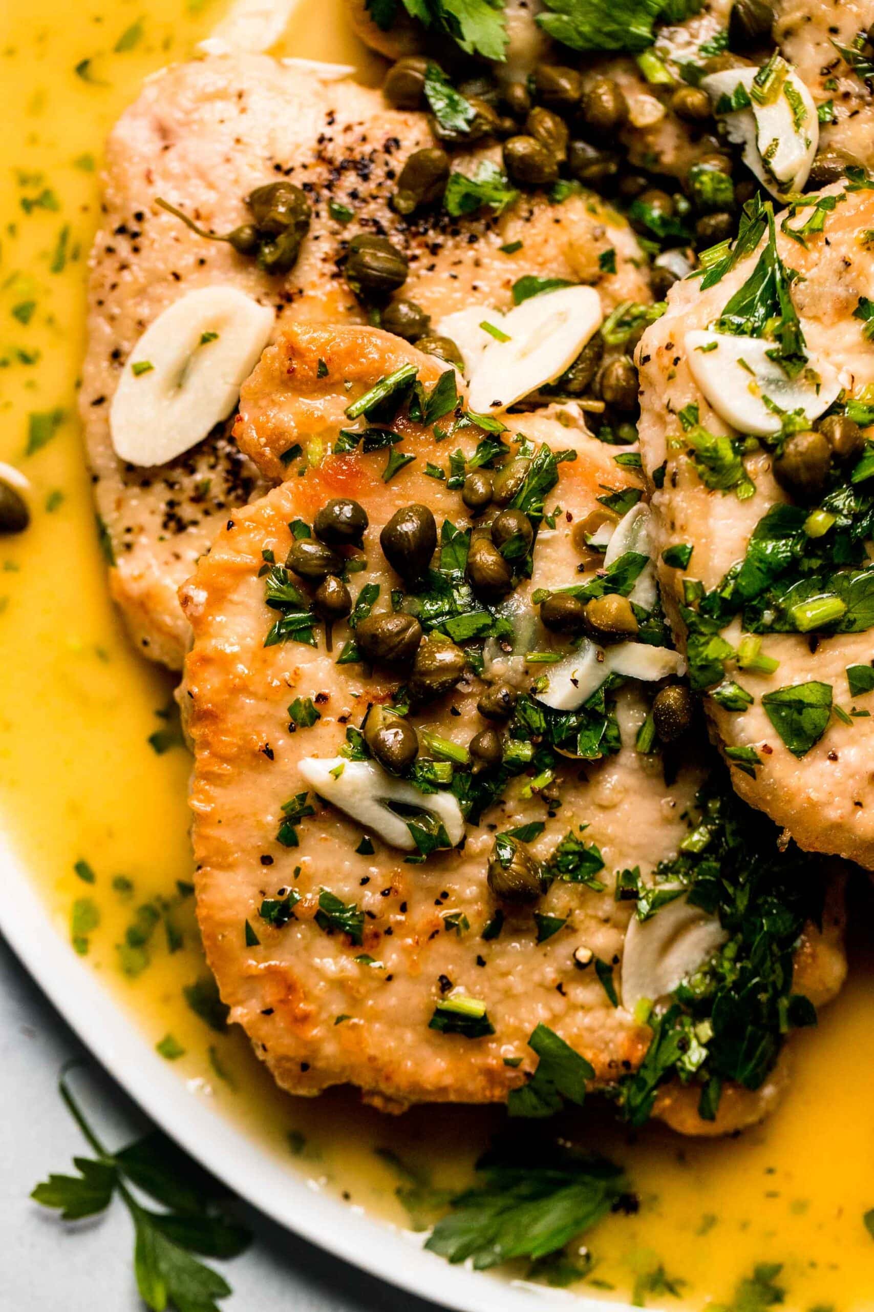 Overhead closeup of pork piccata slices on plate with capers and garlic.