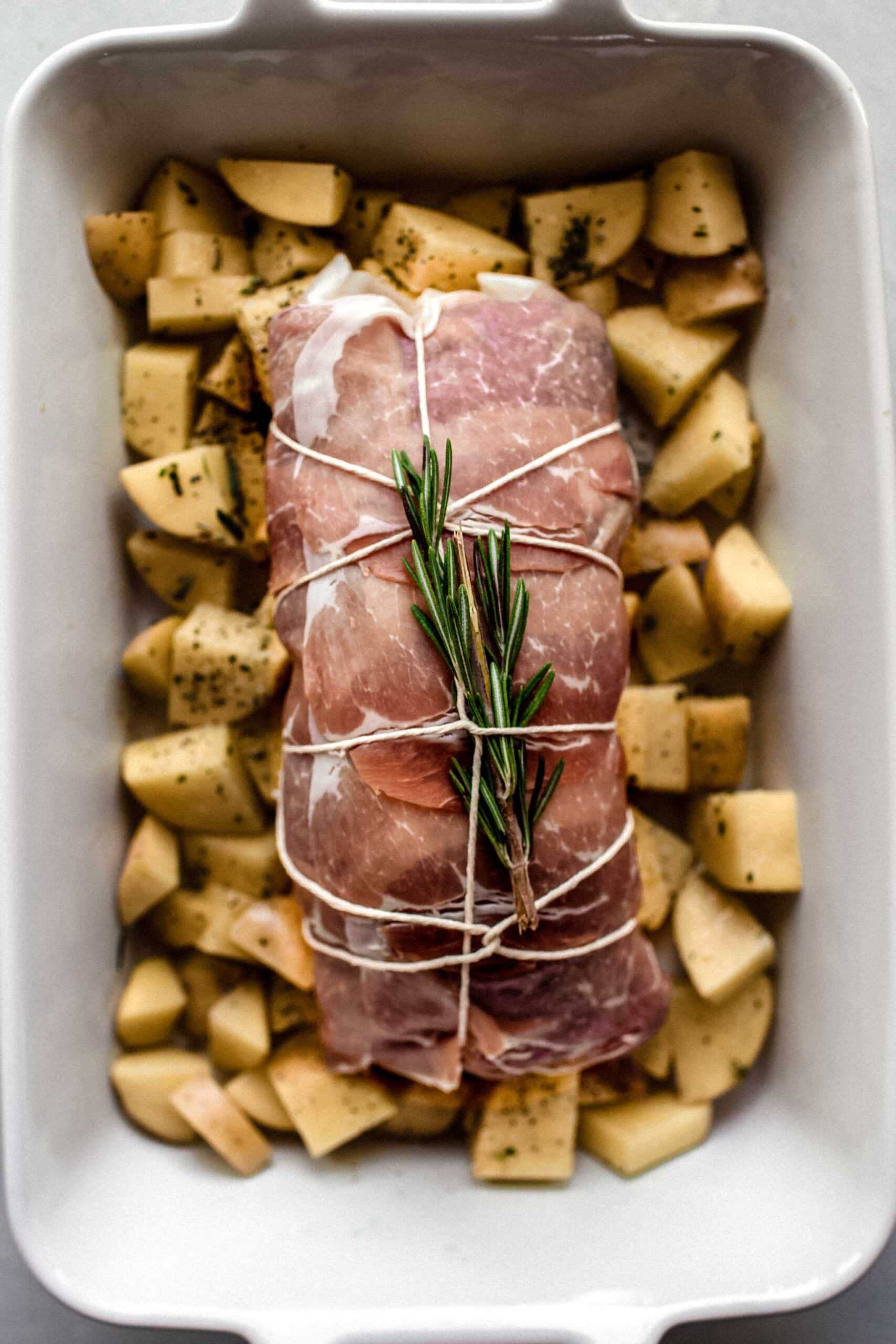 Pork tenderloin wrapped in prosciutto on pan of potatoes before baking.