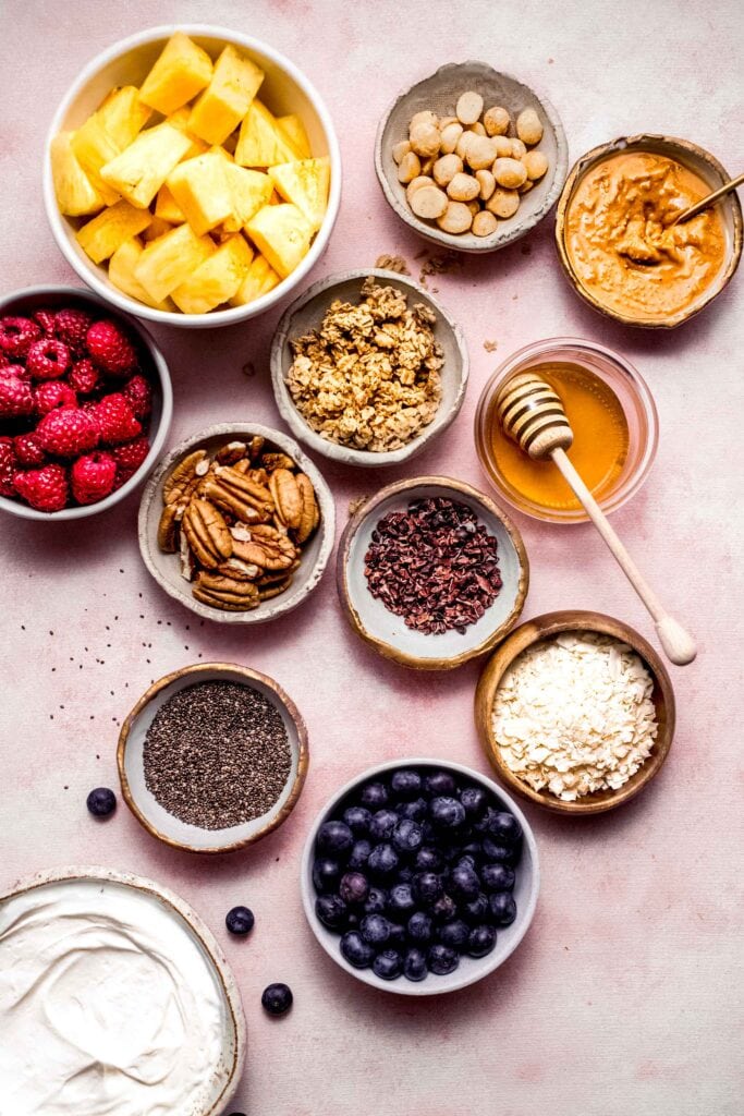 Toppings for greek yogurt parfaits in small bowls on pink counter.