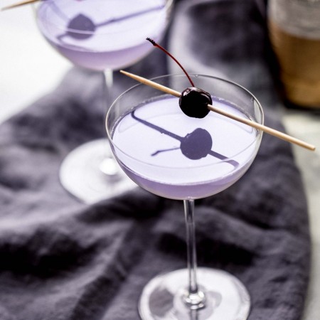 Two aviation cocktails garnished with cherries next to alcohol bottles.
