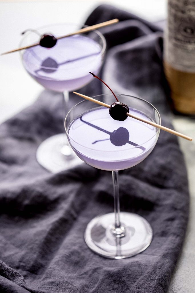 Two aviation cocktails garnished with cherries next to alcohol bottles.