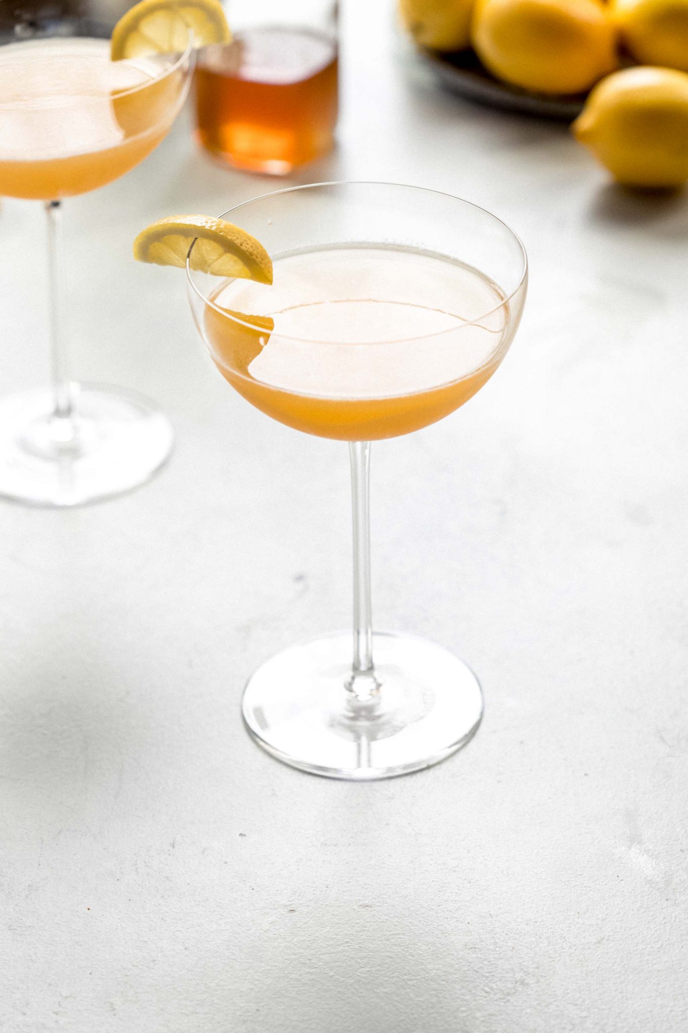 Two Bees Knees Cocktails in coupe glasses next to honey simple syrup and bowl of lemons.