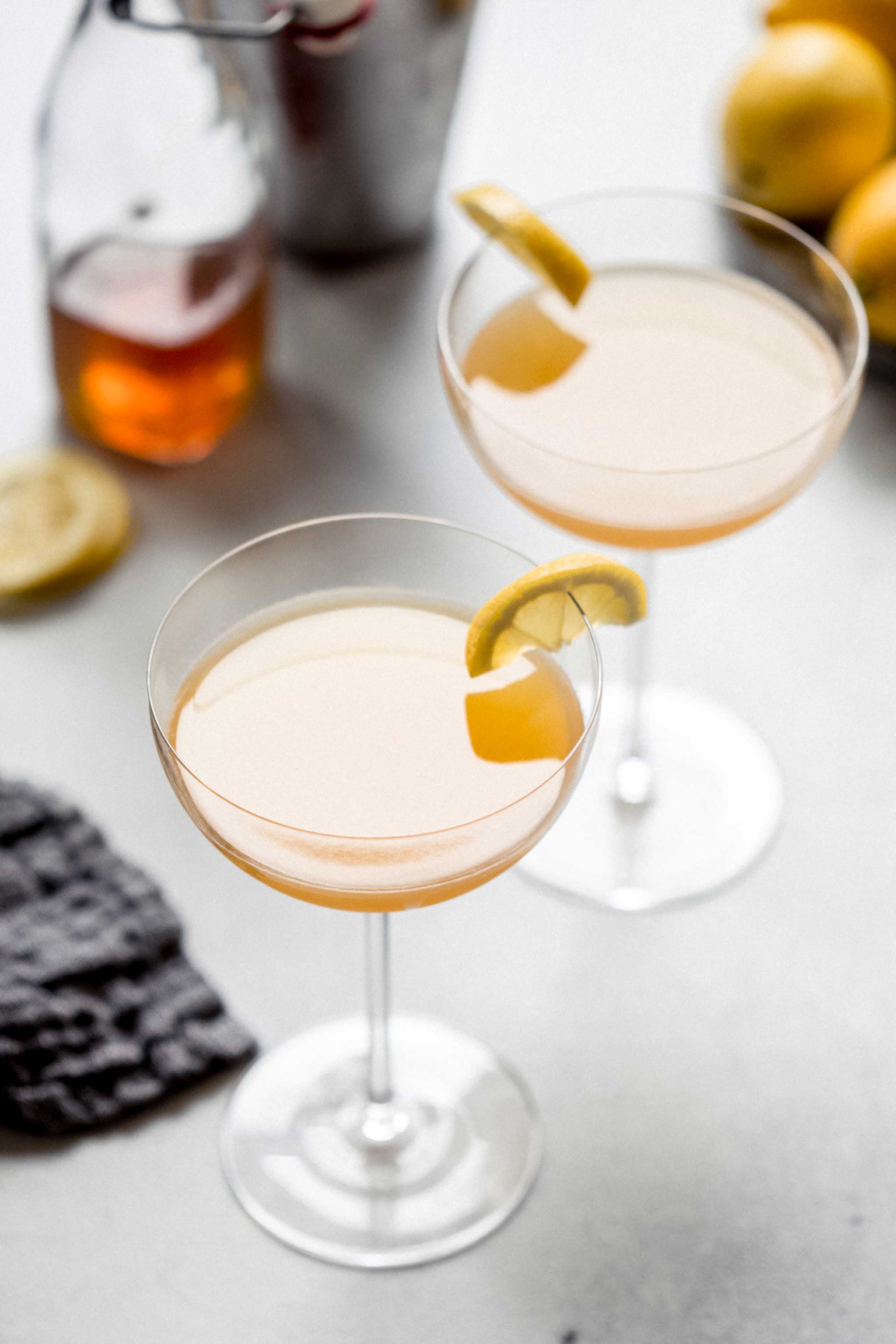 Two Bees Knees Cocktails in coupe glasses next to honey simple syrup and bowl of lemons.