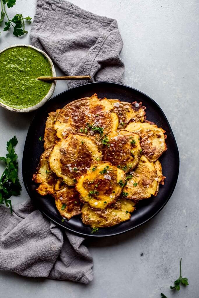 Crispy smashed potatoes on black plate sprinkled with coarse salt next to bowl of mojo sauce.