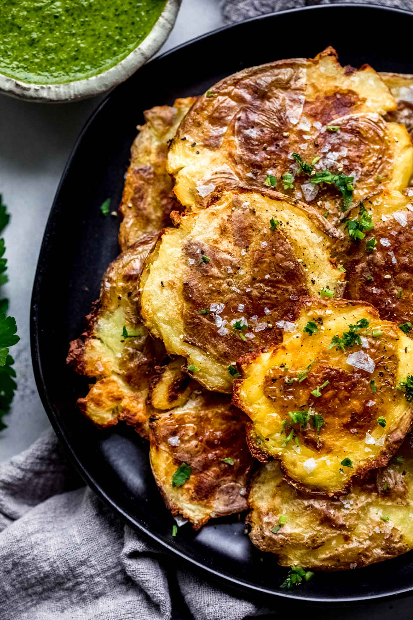 Crispy smashed potatoes on black plate sprinkled with coarse salt next to bowl of mojo sauce.