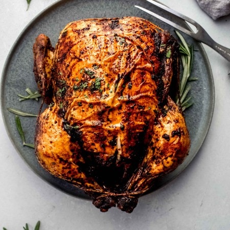 Roasted chicken on grey serving plate with sprig of rosemary and carving fork.