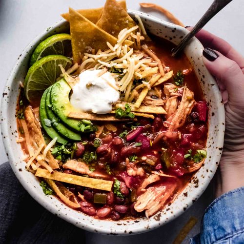 Hand holding bowl of Instant Pot Chicken Tortilla Soup topped with avocado, tortilla chips and sour cream.