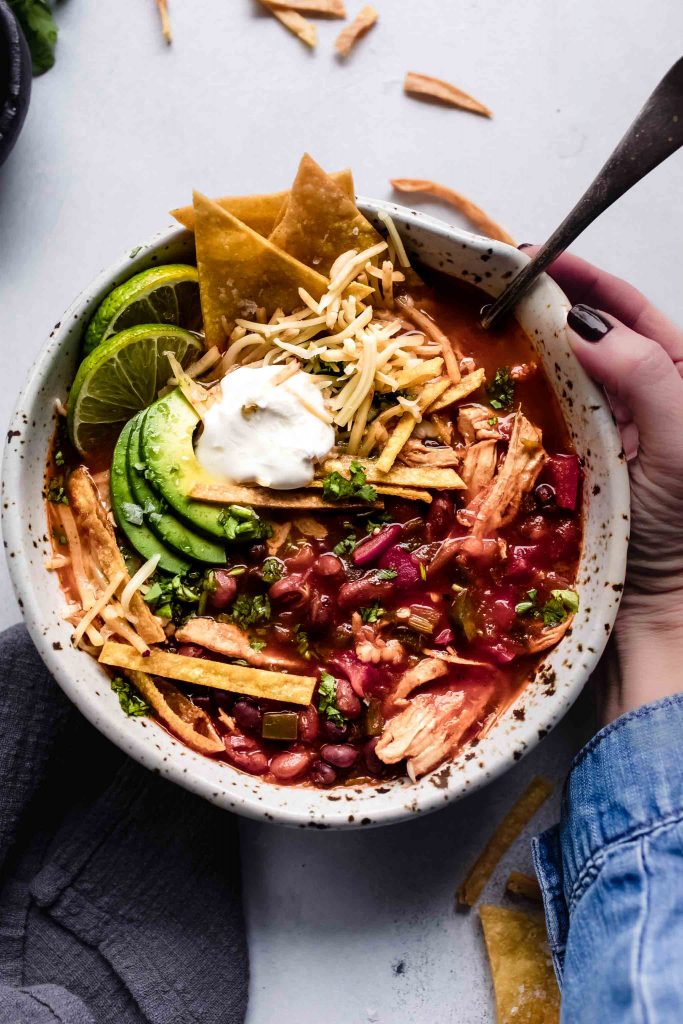 Hand holding bowl of Instant Pot Chicken Tortilla Soup topped with avocado, tortilla chips and sour cream.