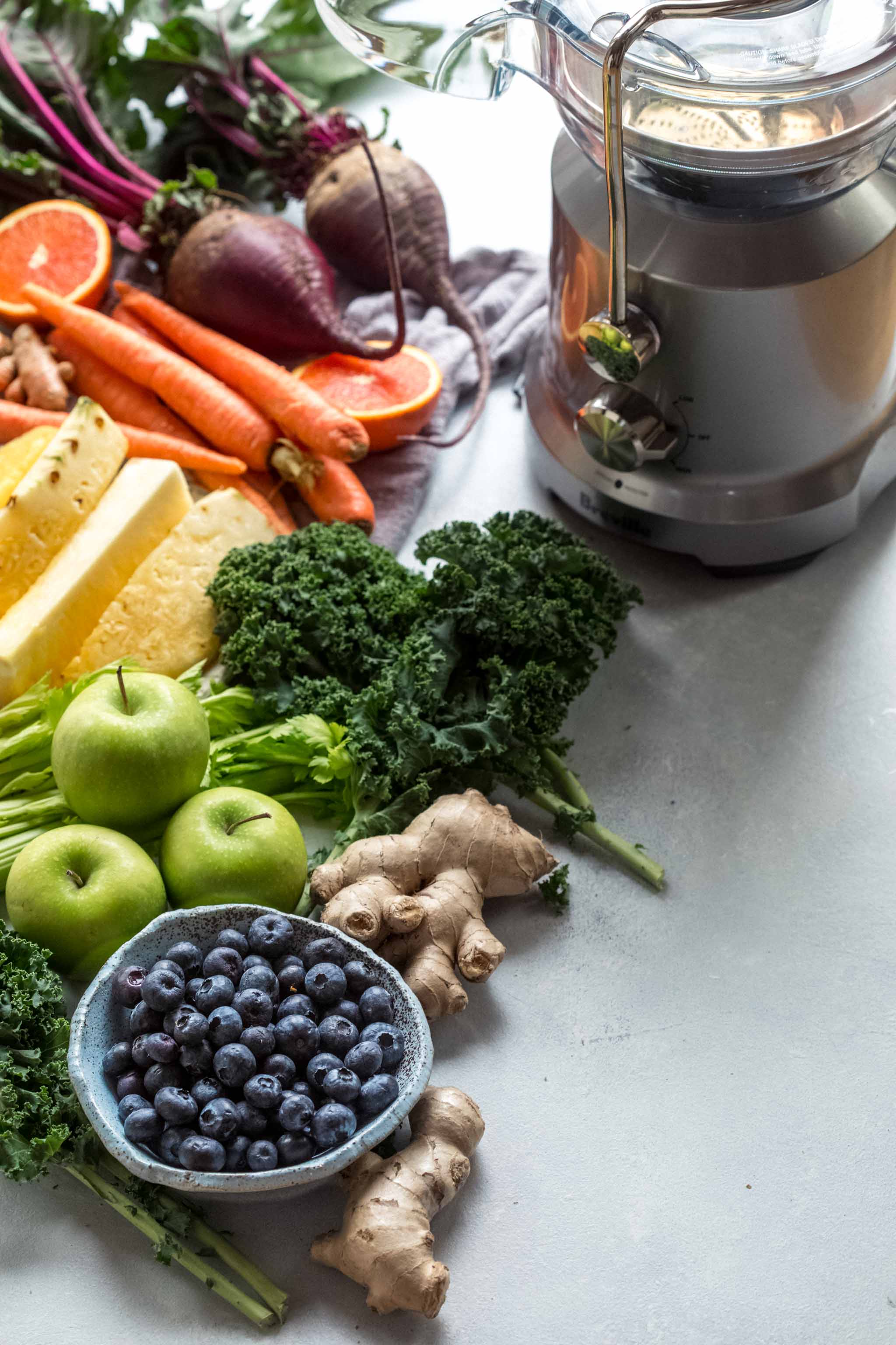 Ingredients for juice cleanse recipes arranged like a rainbow next to juicer.