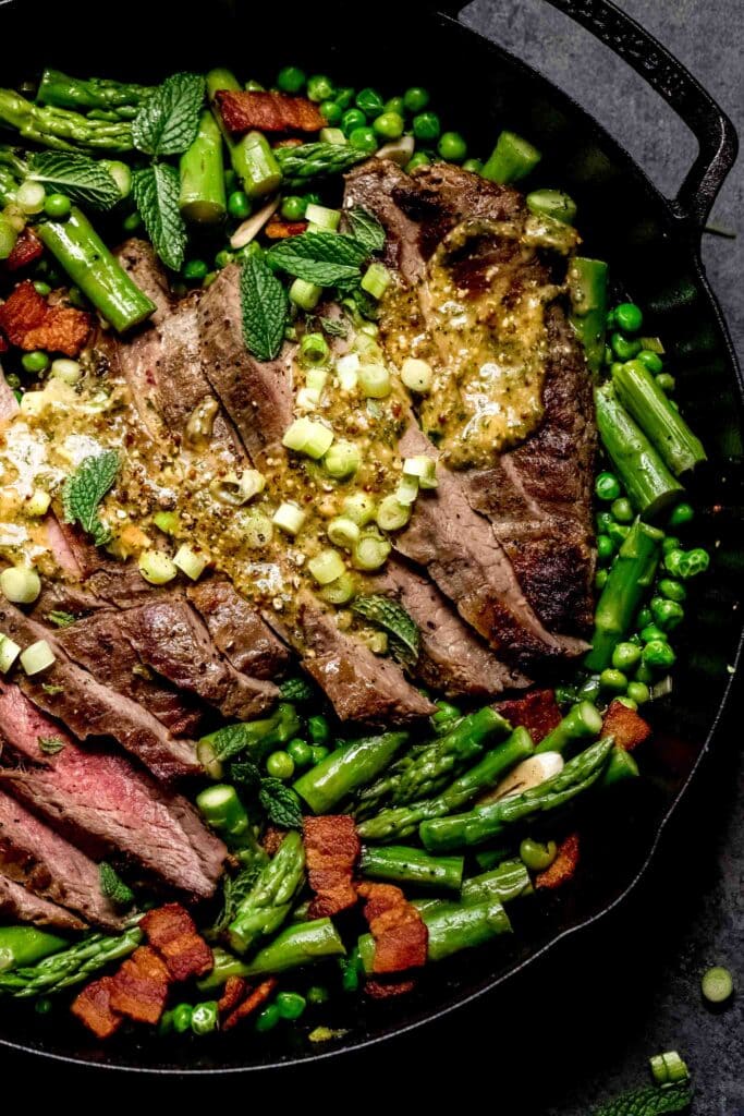 Sliced steak in skillet with asparagus and peas, drizzled with sauce.