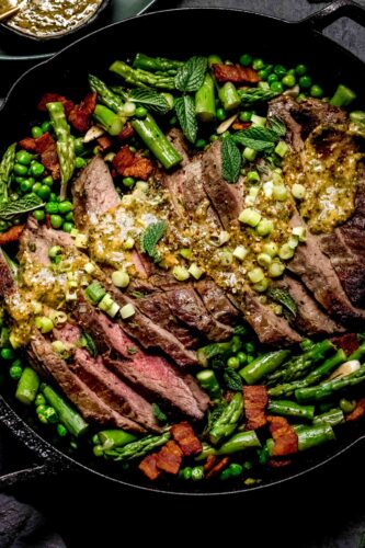 Sliced steak in skillet topped with mustard sauce and surrounded by asparagus and peas.