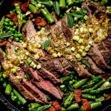 Sliced steak in skillet topped with mustard sauce and surrounded by asparagus and peas.