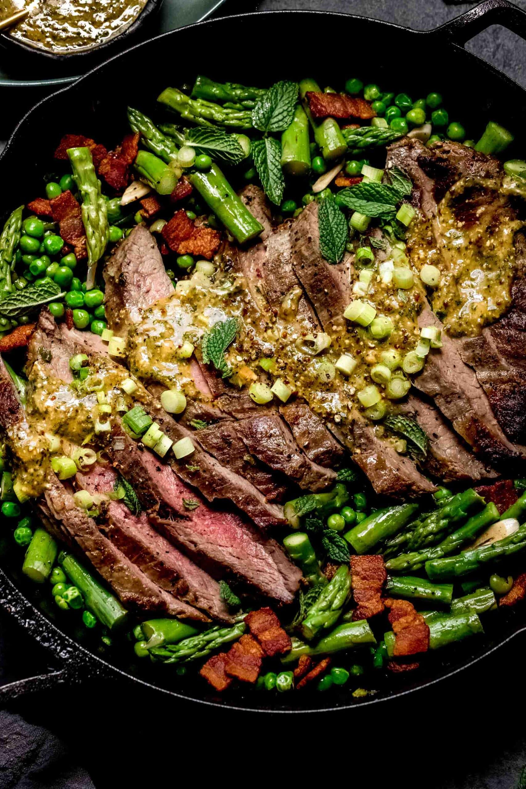 Sliced steak in skillet topped with mustard sauce and surrounded by asparagus and peas.