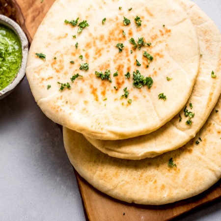Overhead shot of four pieces of naan stacked on wooden cutting board next to bowl of green chutney.