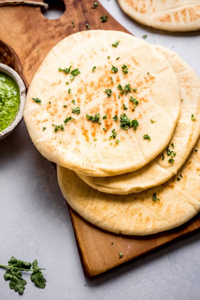 Overhead shot of four pieces of naan stacked on wooden cutting board next to bowl of green chutney.