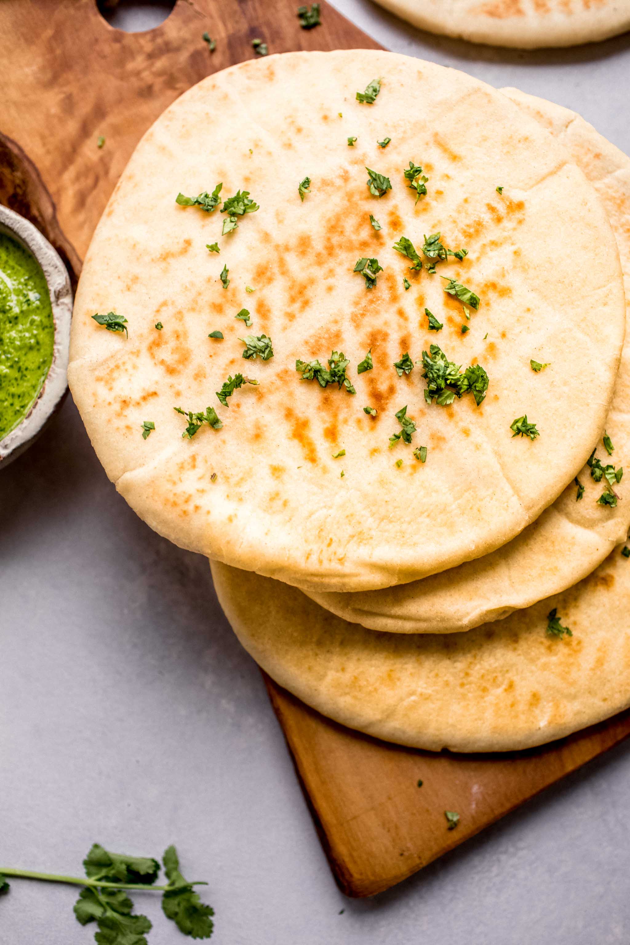 Overhead shot of four pieces of naan stacked on wooden cutting board next to bowl of green chutney.