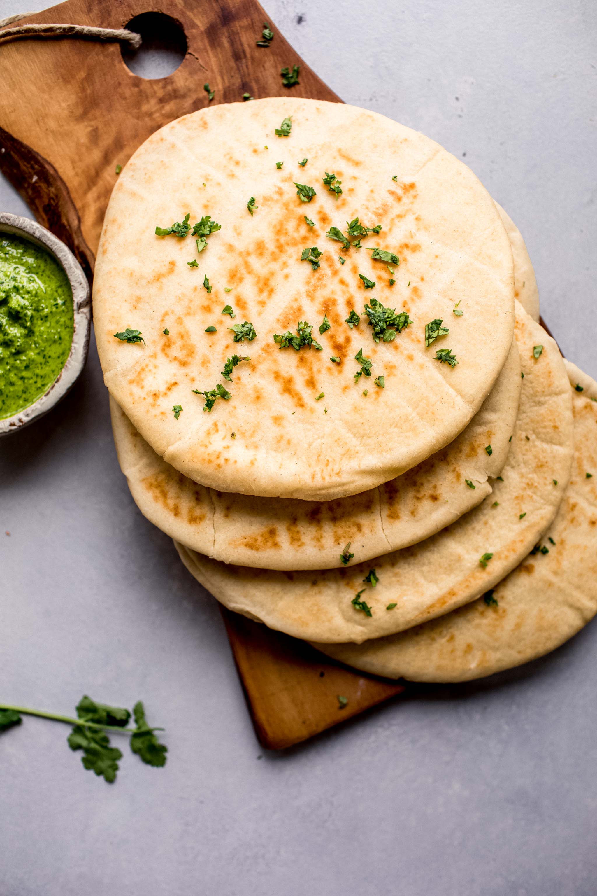 Overhead shot of four pieces of naan stacked on wooden cutting board next to bowl of green chutney. 