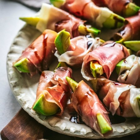 Side view of Avocado bites wrapped in prosciutto on wood cutting board.
