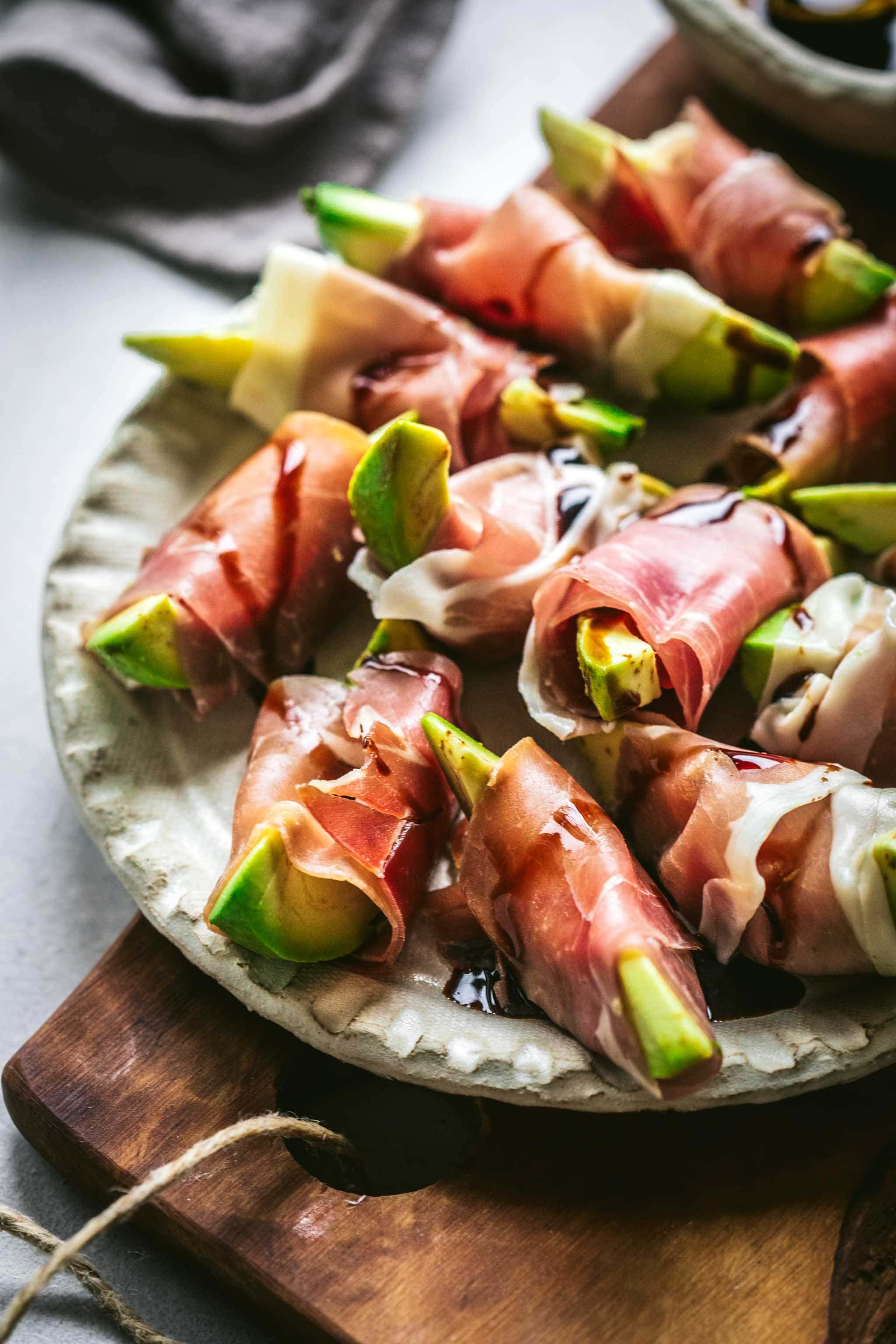 Side view of Avocado bites wrapped in prosciutto on wood cutting board.