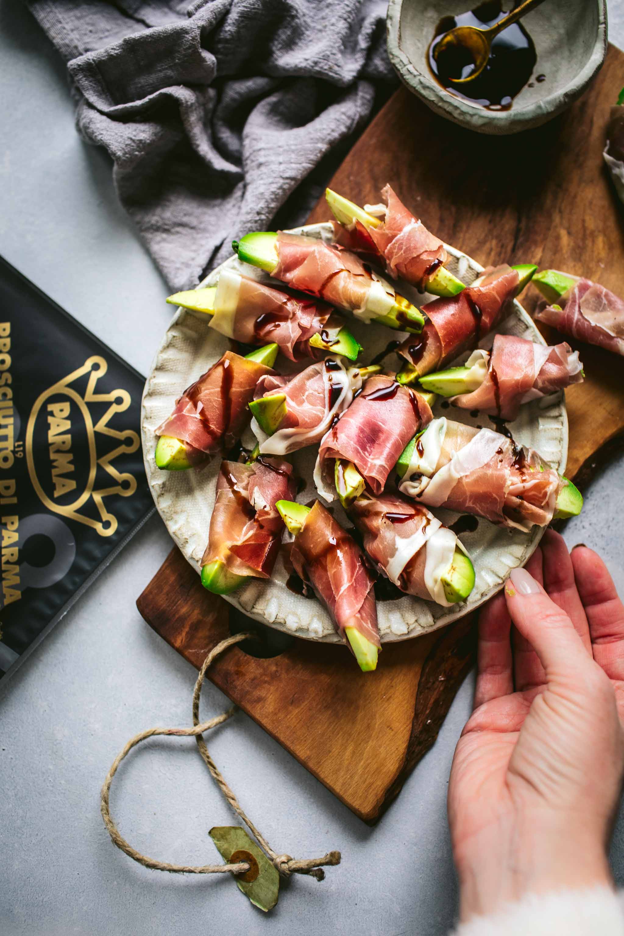 Hand holding plate of Avocado bites wrapped in prosciutto on wood cutting board.
