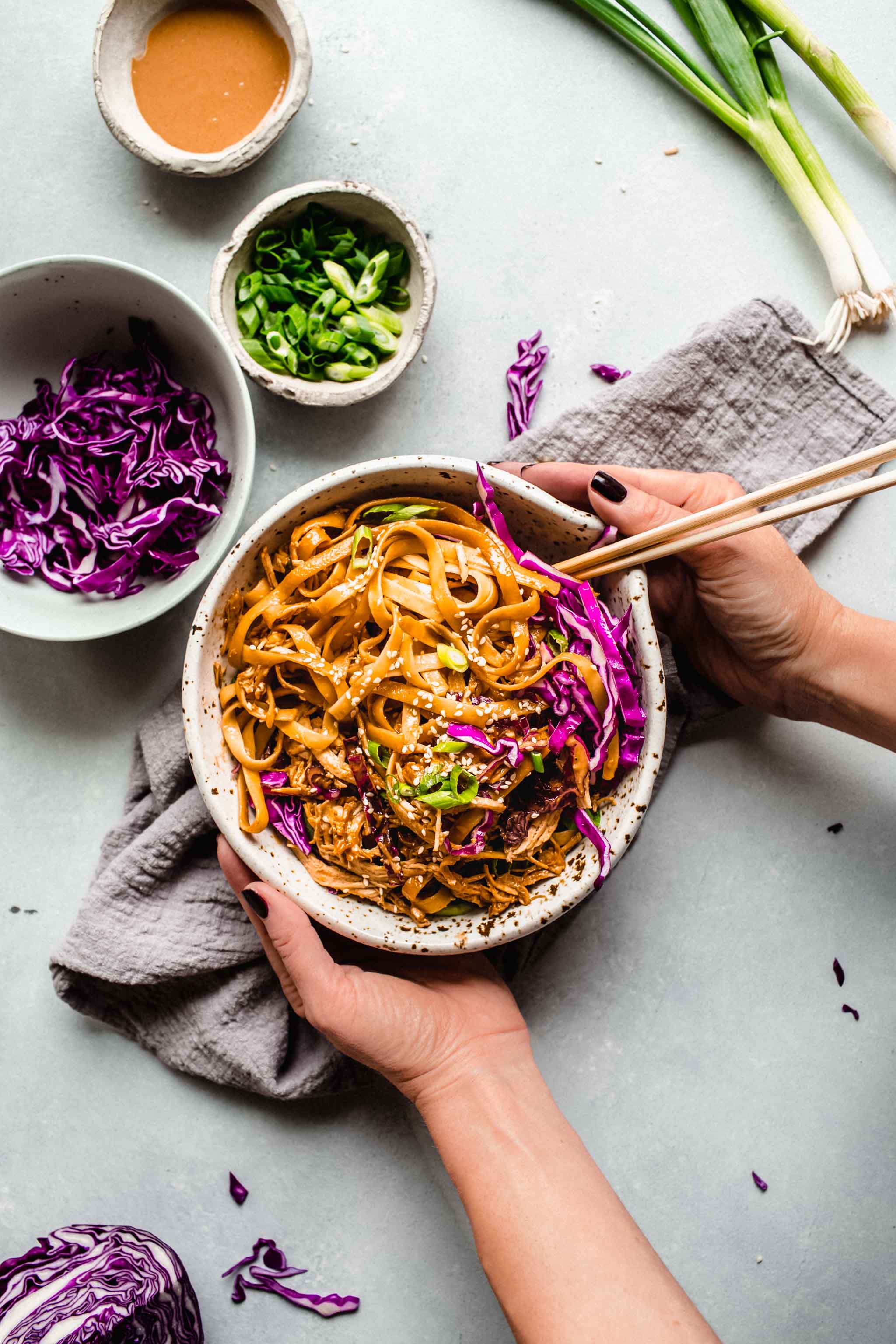 Two hands with chopsticks holding bowl of sesame noodles with chopsticks.