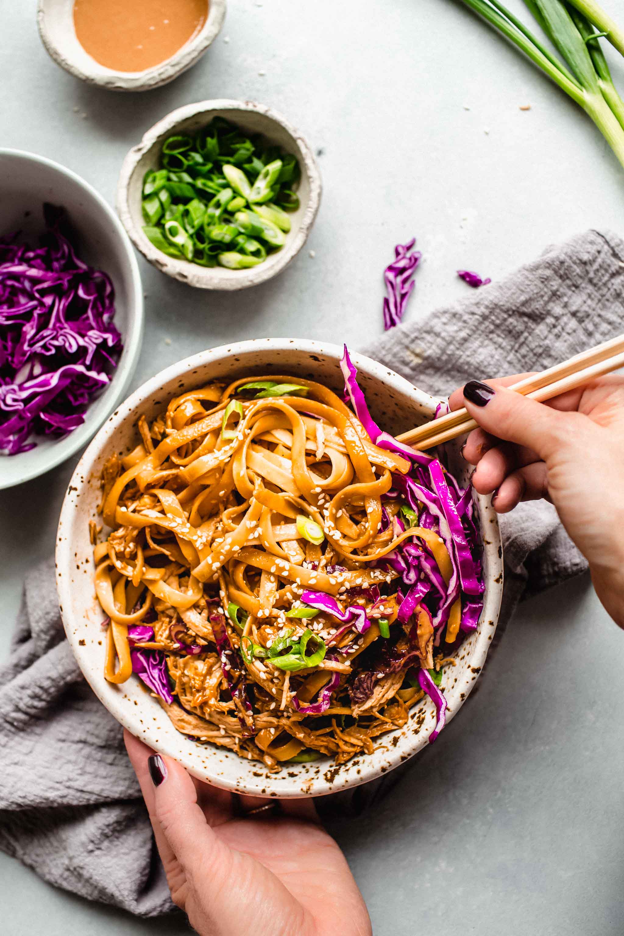 Two hands with chopsticks holding bowl of sesame chicken noodles with chopsticks.