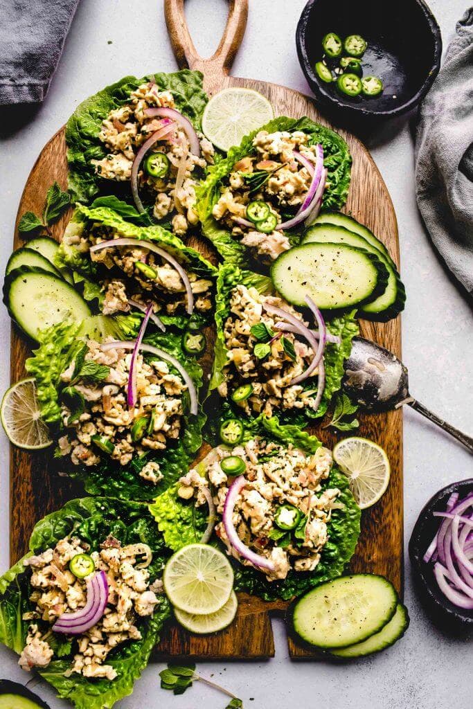 Larb gai lettuce wraps arranged on wood serving tray garnished with red onions and cucumber slices.