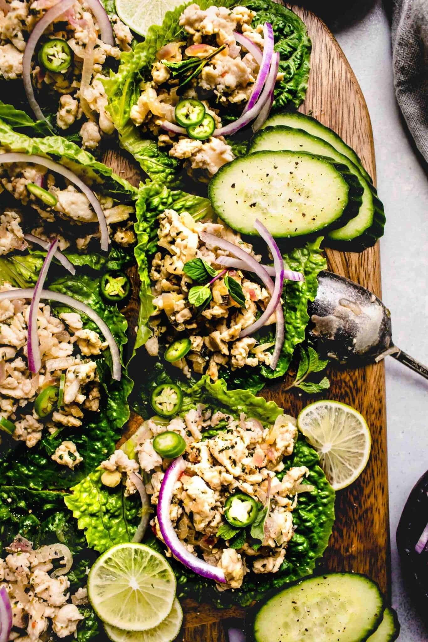 Larb gai lettuce wraps arranged on wood serving tray garnished with red onions and cucumber slices.