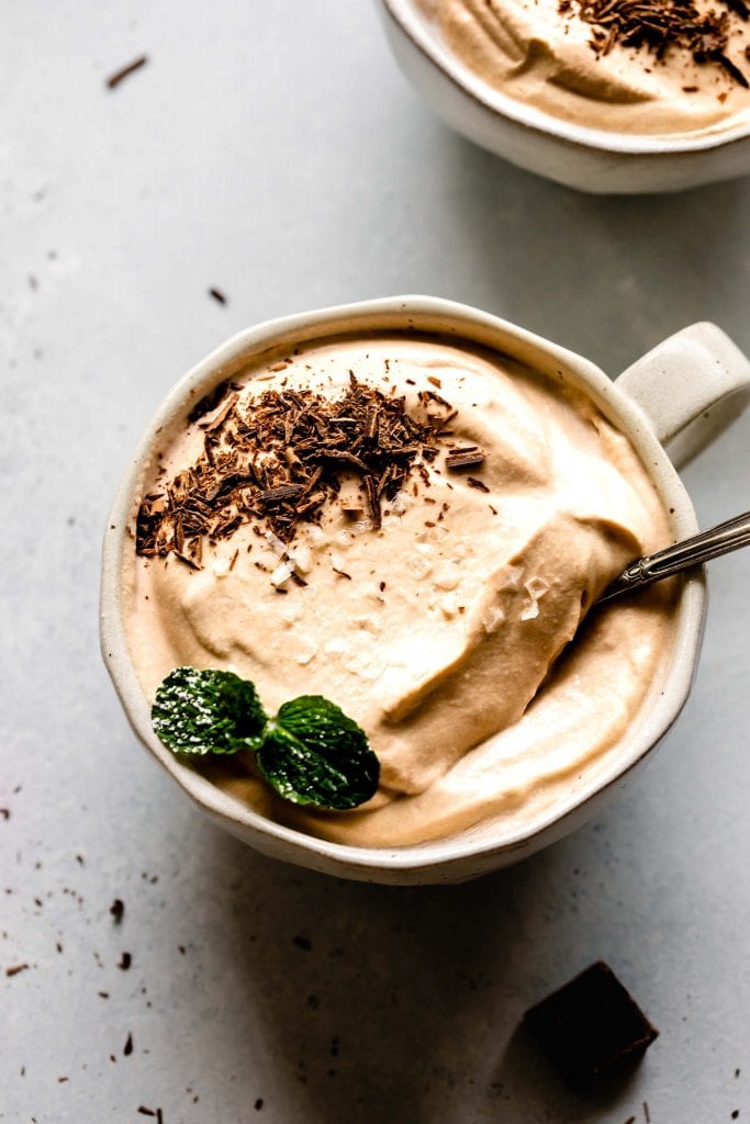 Overhead close up of bowl of coffee mousse topped with flaky salt, mint sprig and chocolate shavings.