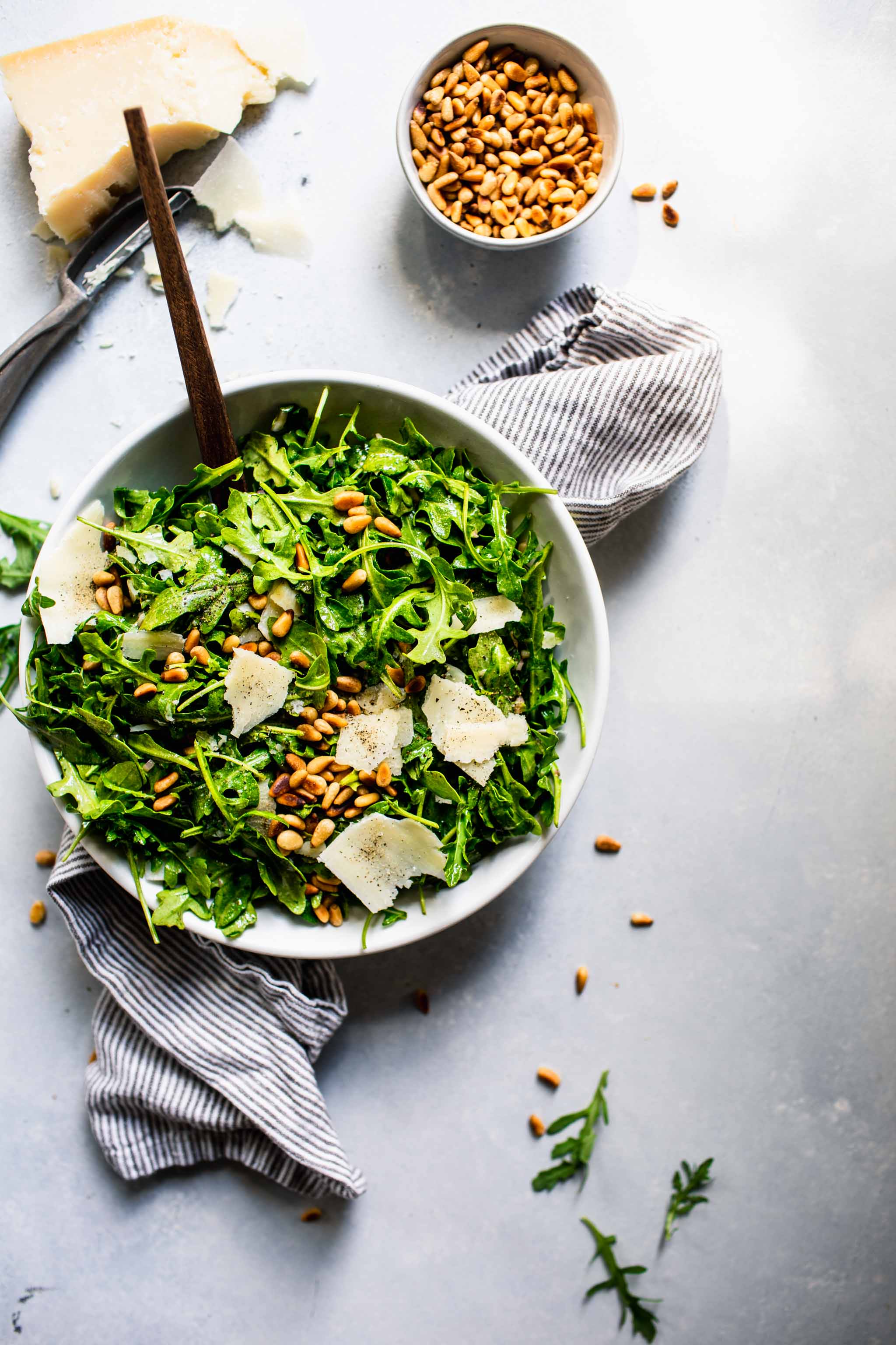 Overhead shot of arugula salad next to shaved parmesan and bowl of pine nuts.