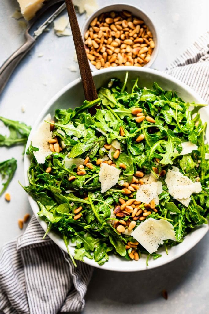 Overhead shot of arugula salad next to bowl of pine nuts and shaved parmesan.