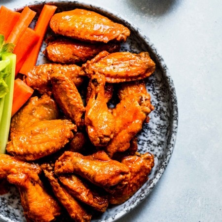 Baked buffalo wings coated in buffalo sauce on grey speckled plate next to blue cheese and celery sticks.