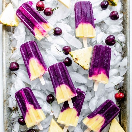Cherry pineapple popsicles on tray of ice cubes with pineapple wedges and cherries scattered about.
