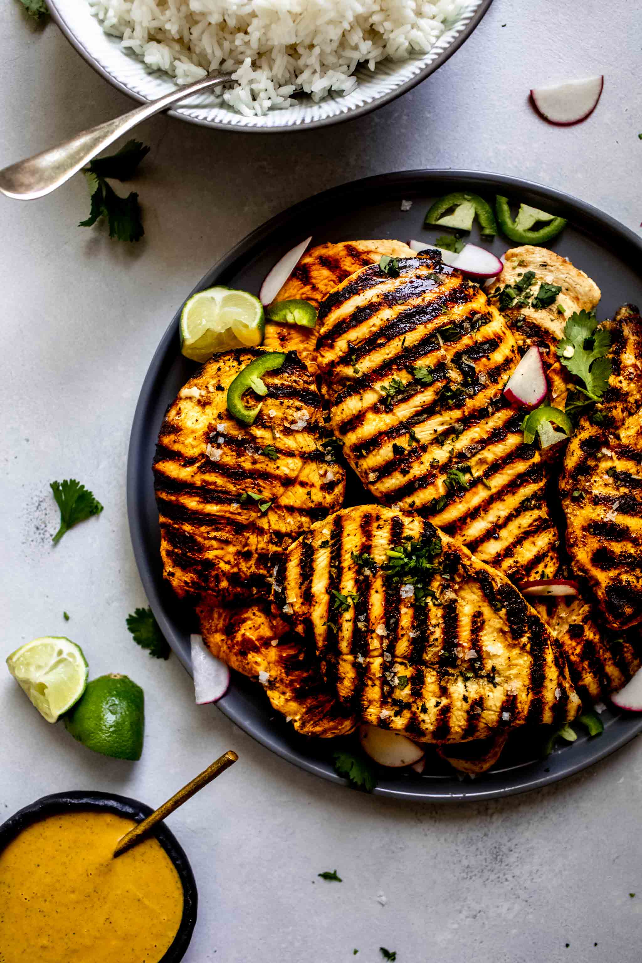 Grilled chicken breasts on platter next to bowl of peanut sauce and bowl of rice.