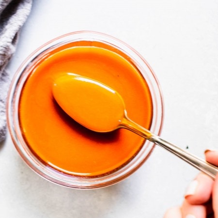 Hand holding spoon dipped into jar of buffalo sauce.