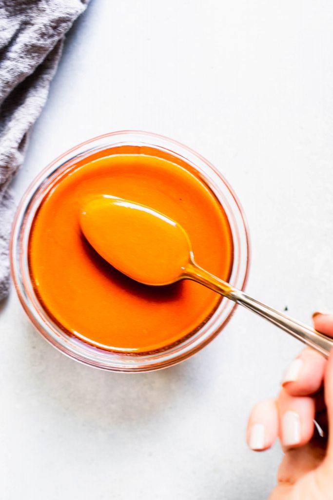 Hand holding spoon dipped into jar of buffalo sauce.