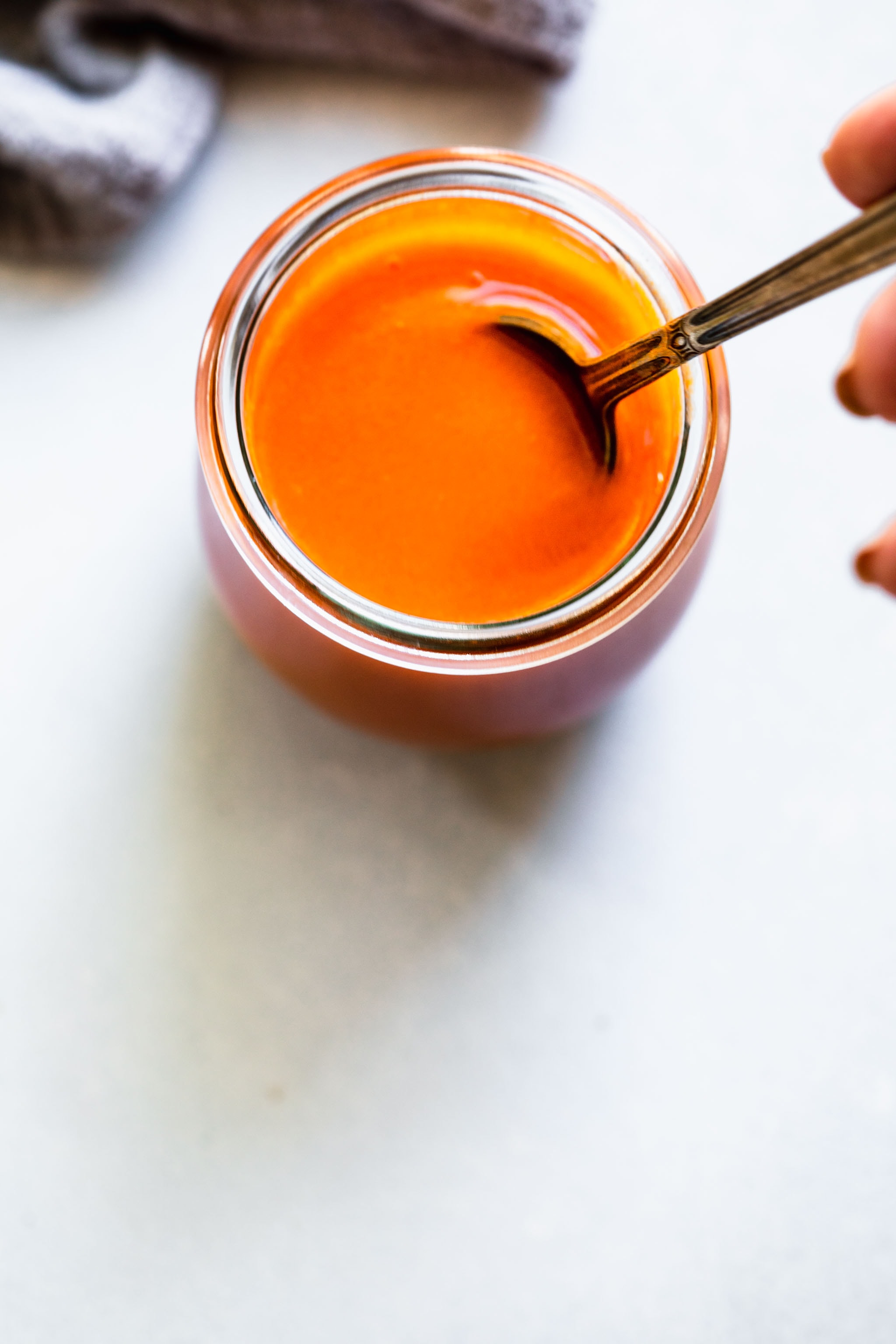 Hand holding spoon dipped into jar of buffalo sauce.