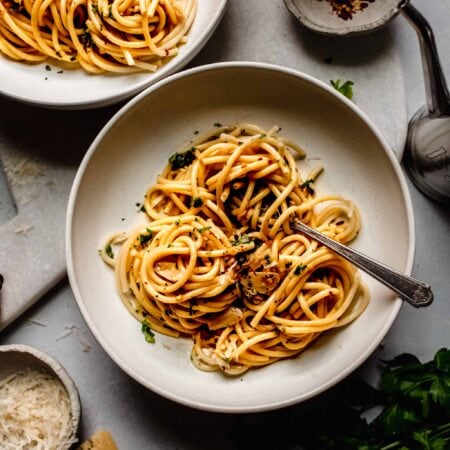 Overhead shot of two bowls of garlic and oil pasta next to small bowls of chili flakes and parmesan.