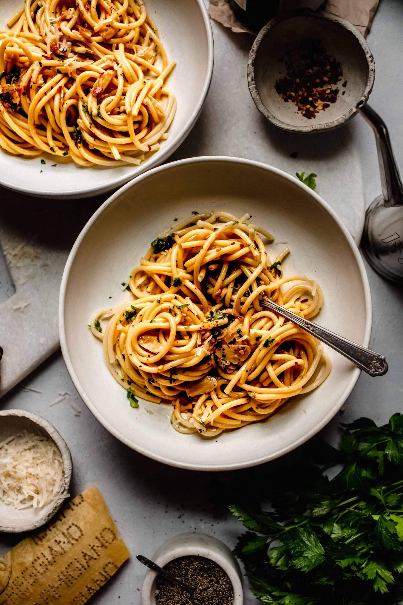 Overhead shot of two bowls of garlic and oil pasta next to small bowls of chili flakes and parmesan.