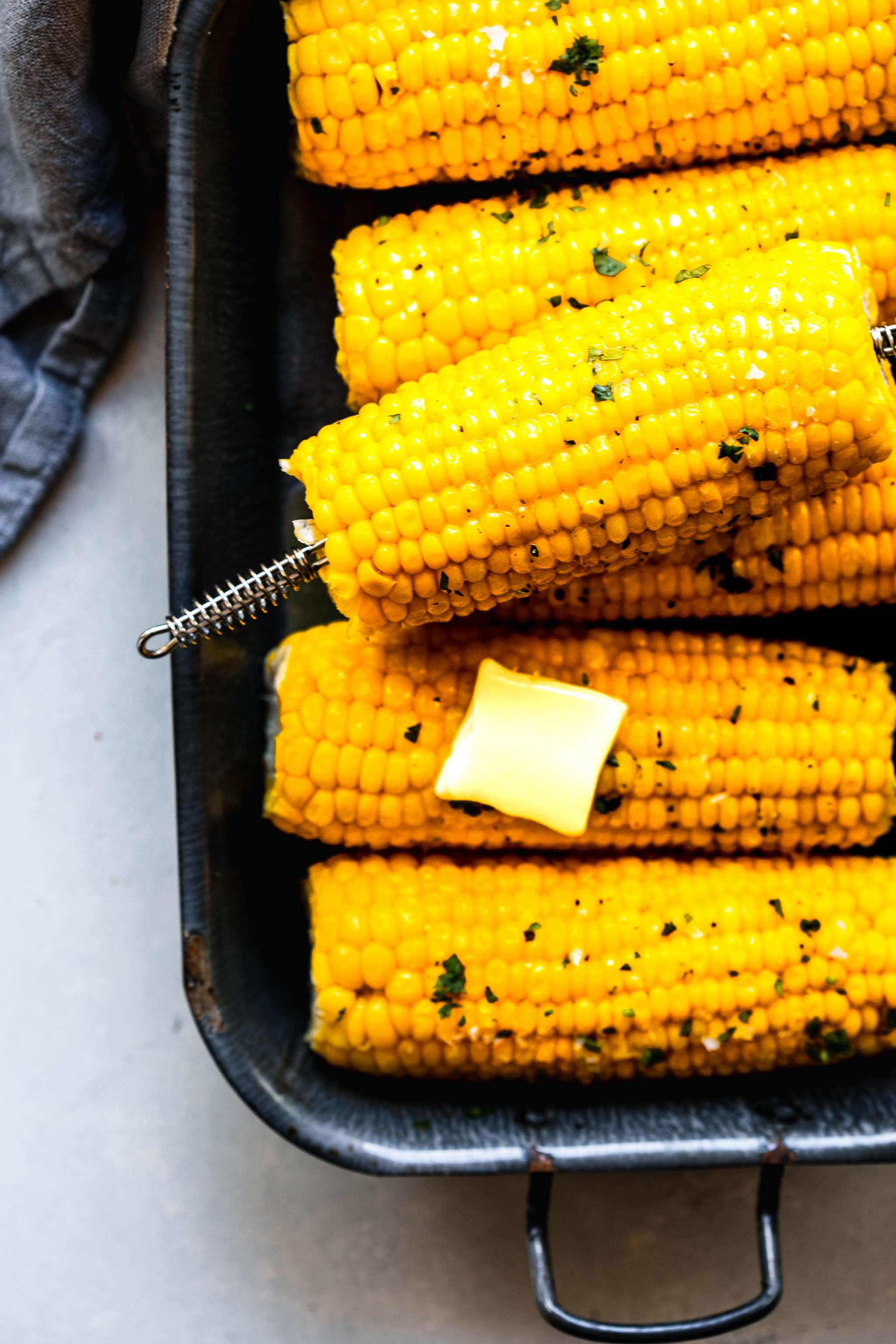 Ears of corn on the cob in a grey serving dish with a pat of butter and sprinkling of parsley.